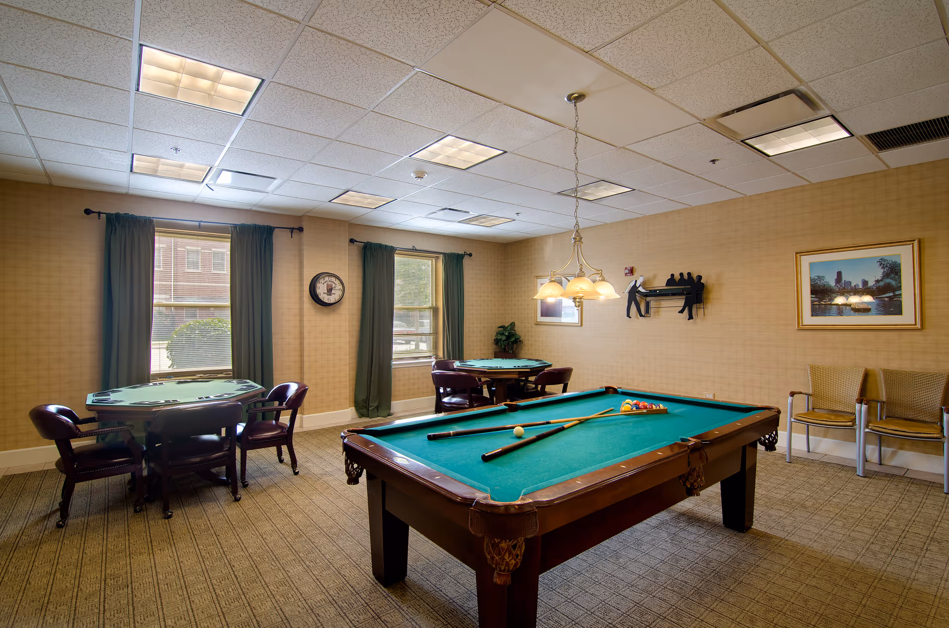 A recreational room with a pool table in the center, two card tables with chairs near windows with green curtains, a wall clock, framed pictures, and a wall decoration of two people playing pool. The room has beige walls and carpeted floor with overhead fluorescent lighting.