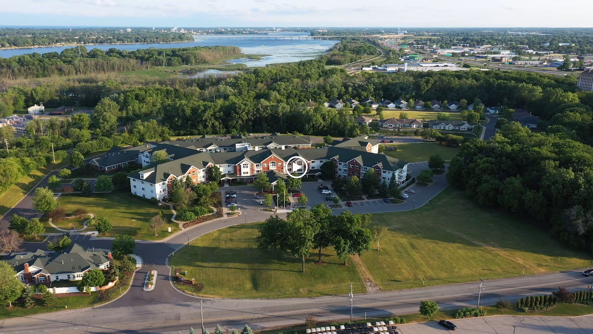 Aerial view of the Touchmark on West Prospect senior living facility surrounded by green trees and open grassy areas, with a river and bridge visible in the background under a clear sky.