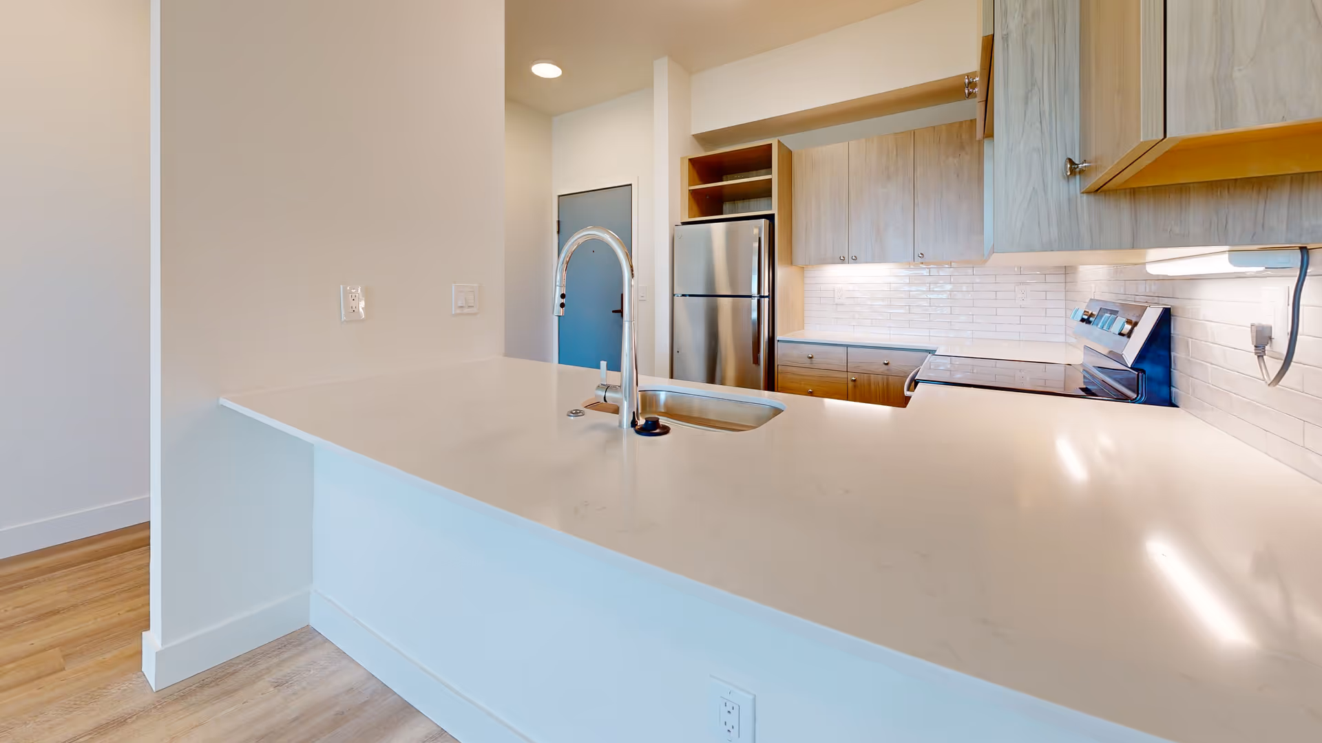 Modern kitchen with a white countertop island featuring a sink and faucet, stainless steel refrigerator and stove, and light wood cabinets.