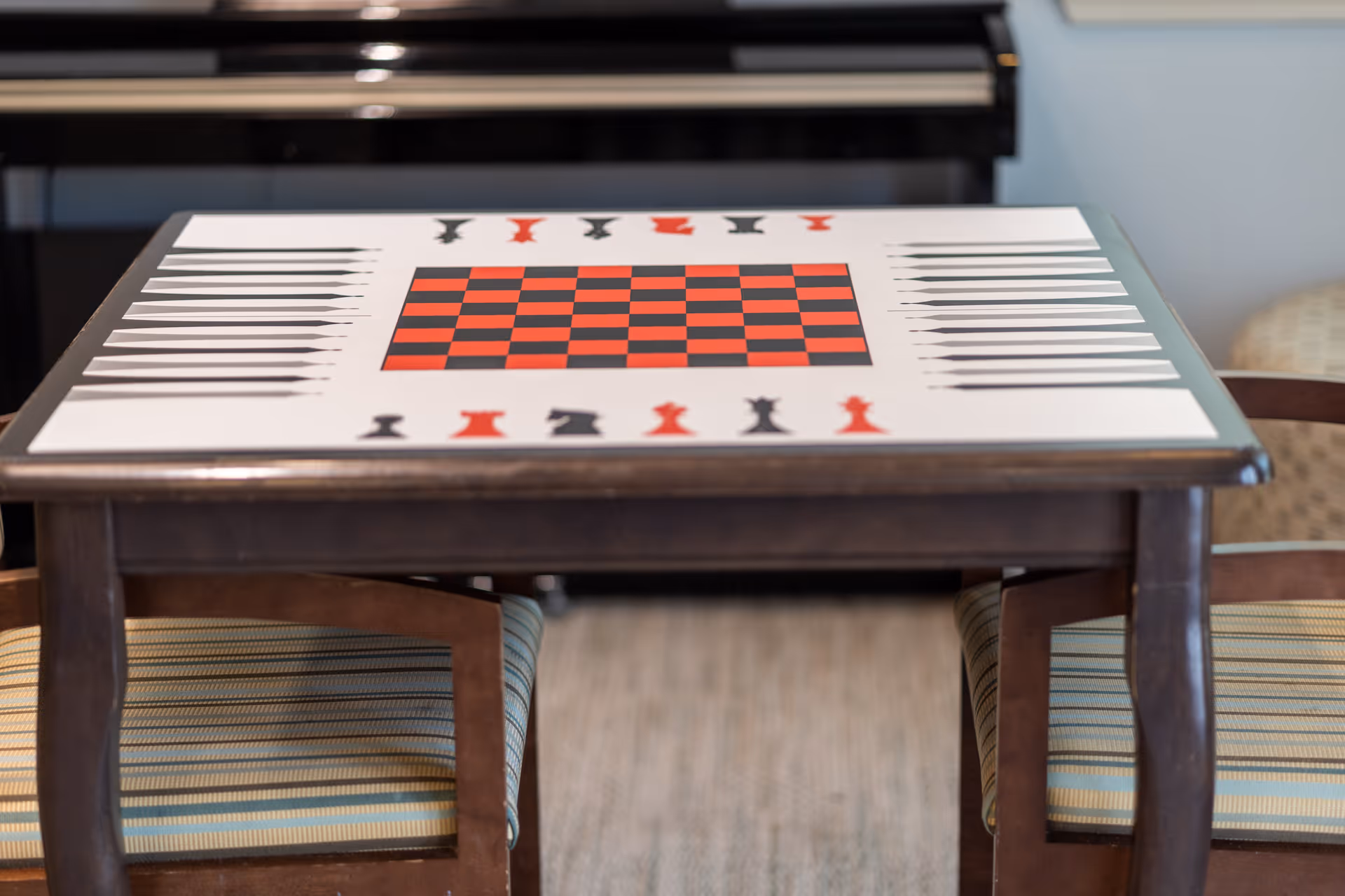 Wooden table with a red-and-black chess/checker board printed on its surface and two striped chairs in a common room.