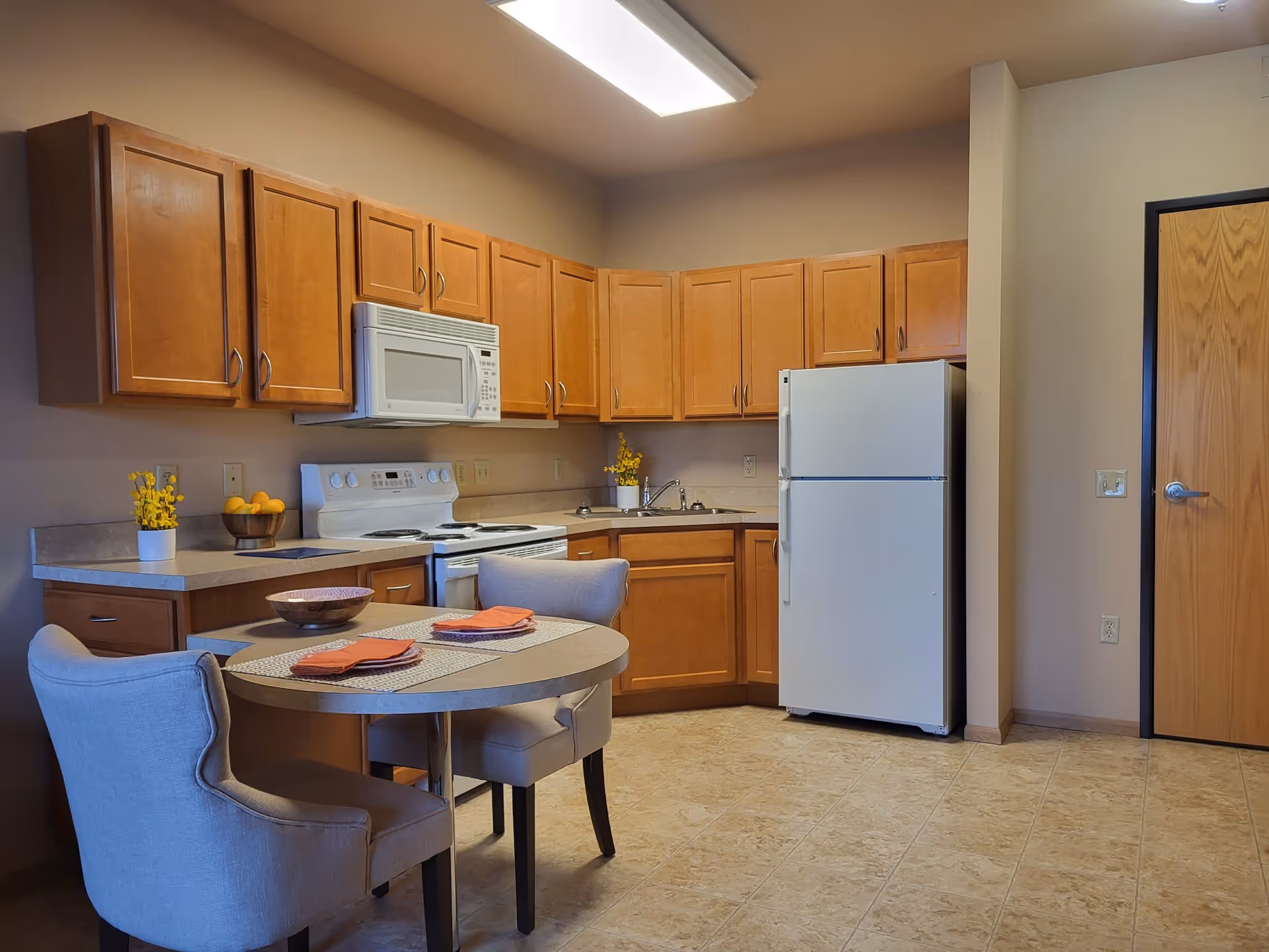 A kitchen area in a senior living apartment featuring wooden cabinets, a white refrigerator, a white stove with an overhead microwave, and a round dining table with two upholstered chairs. The table is set with placemats, orange napkins, and a decorative bowl. The floor is tiled, and there is a wooden door on the right side of the image.