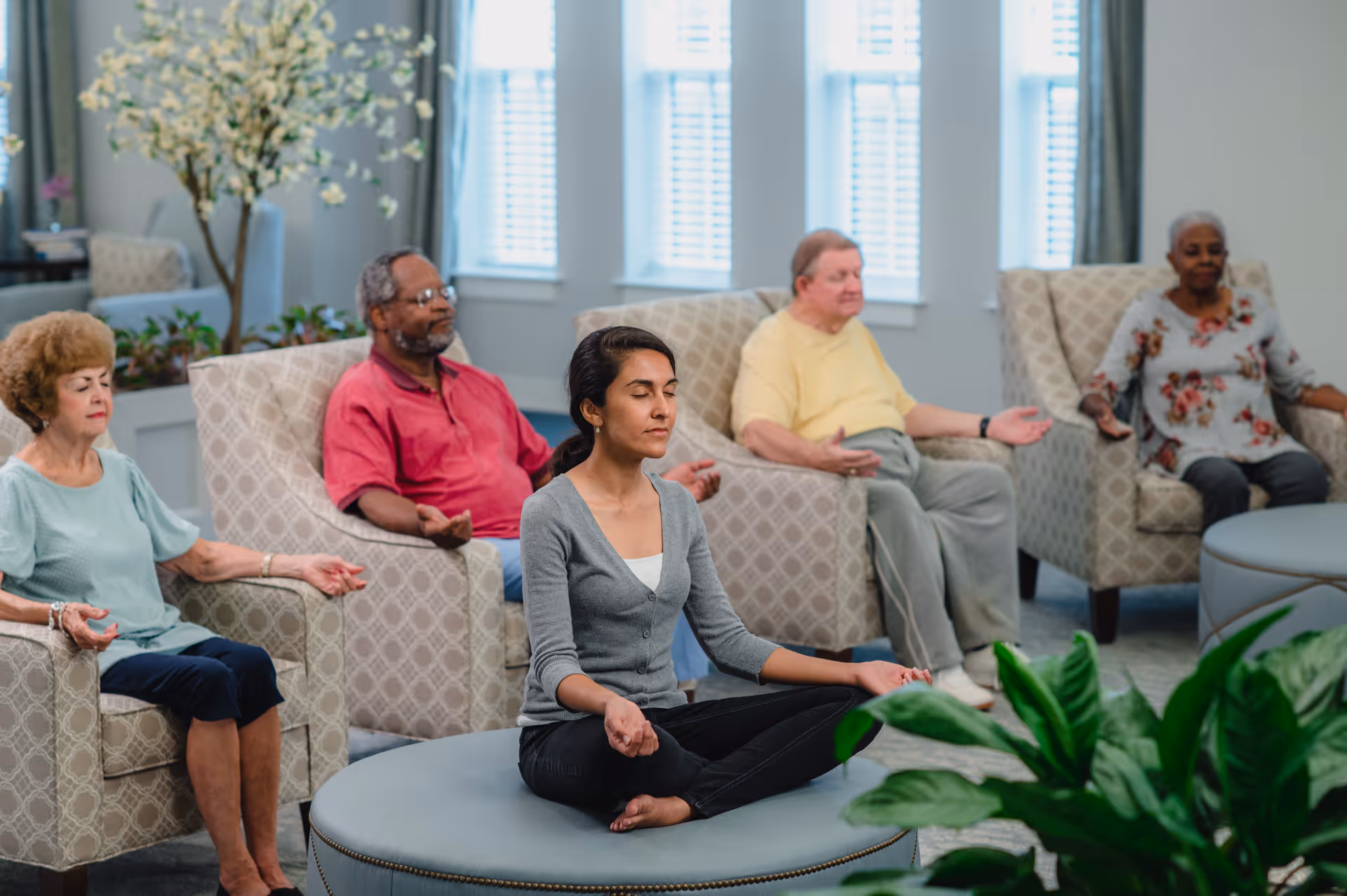 A group of four elderly people and a younger woman sitting in a living room area meditating with their eyes closed. The elderly individuals are seated in armchairs while the younger woman is sitting cross-legged on a round ottoman. The room is bright with large windows and decorated with plants and light-colored furniture.