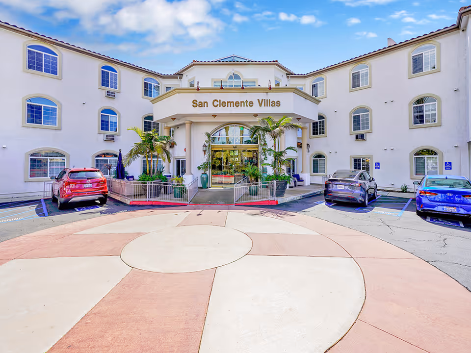 Front exterior view of San Clemente Villas, a three-story building with arched windows and a central entrance featuring glass doors and potted plants. There are several cars parked in front, including in designated handicapped parking spaces. The sky is partly cloudy.