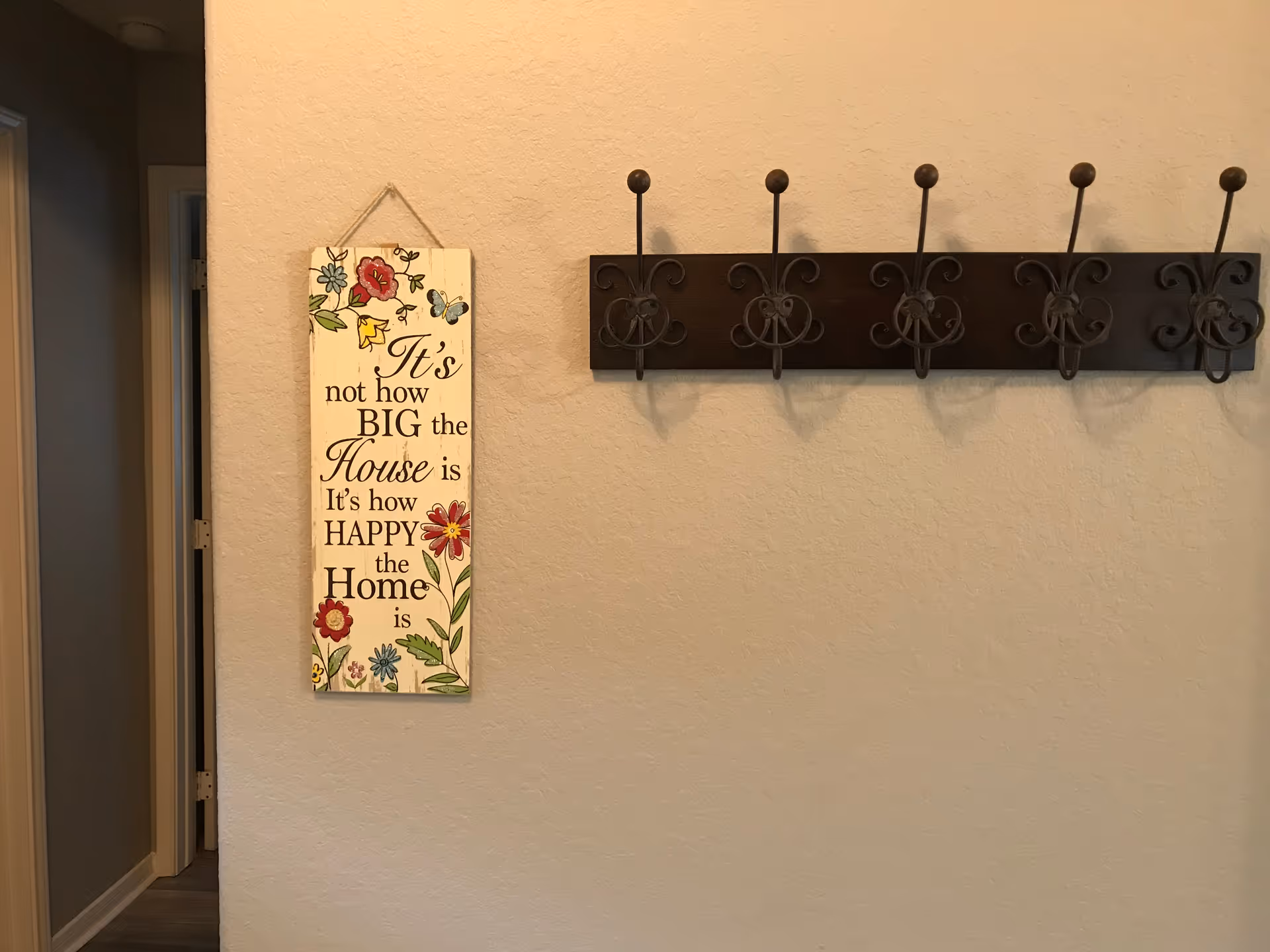 A beige wall with a decorative wooden sign hanging on the left side that reads 'It's not how BIG the House is It's how HAPPY the Home is' with floral and butterfly designs. To the right, there is a dark wooden coat rack with five ornate metal hooks. A doorway is visible on the far left side of the image.