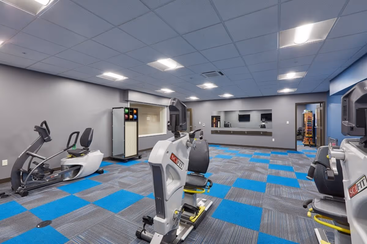 A fitness room with exercise equipment including recumbent bikes and a stepper machine. The room has a gray and blue checkered carpet, gray walls, a large mirror on one wall, and bright overhead lighting. There is a rack with colorful exercise balls and weights in the corner.