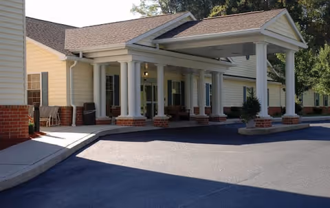 Exterior view of a senior living facility entrance with a covered drop-off area supported by white columns, beige siding, and brick accents. There is a paved driveway and some greenery near the entrance.