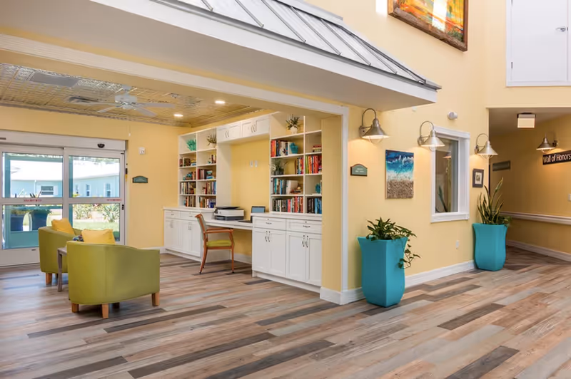 A bright and welcoming interior common area with light yellow walls and wood-patterned flooring. There are two green armchairs with yellow cushions near a large window and glass door. A built-in white bookshelf and desk area with books and a printer is visible. Two large turquoise planters with green plants are placed against the walls. Wall-mounted lights and framed artwork decorate the space.