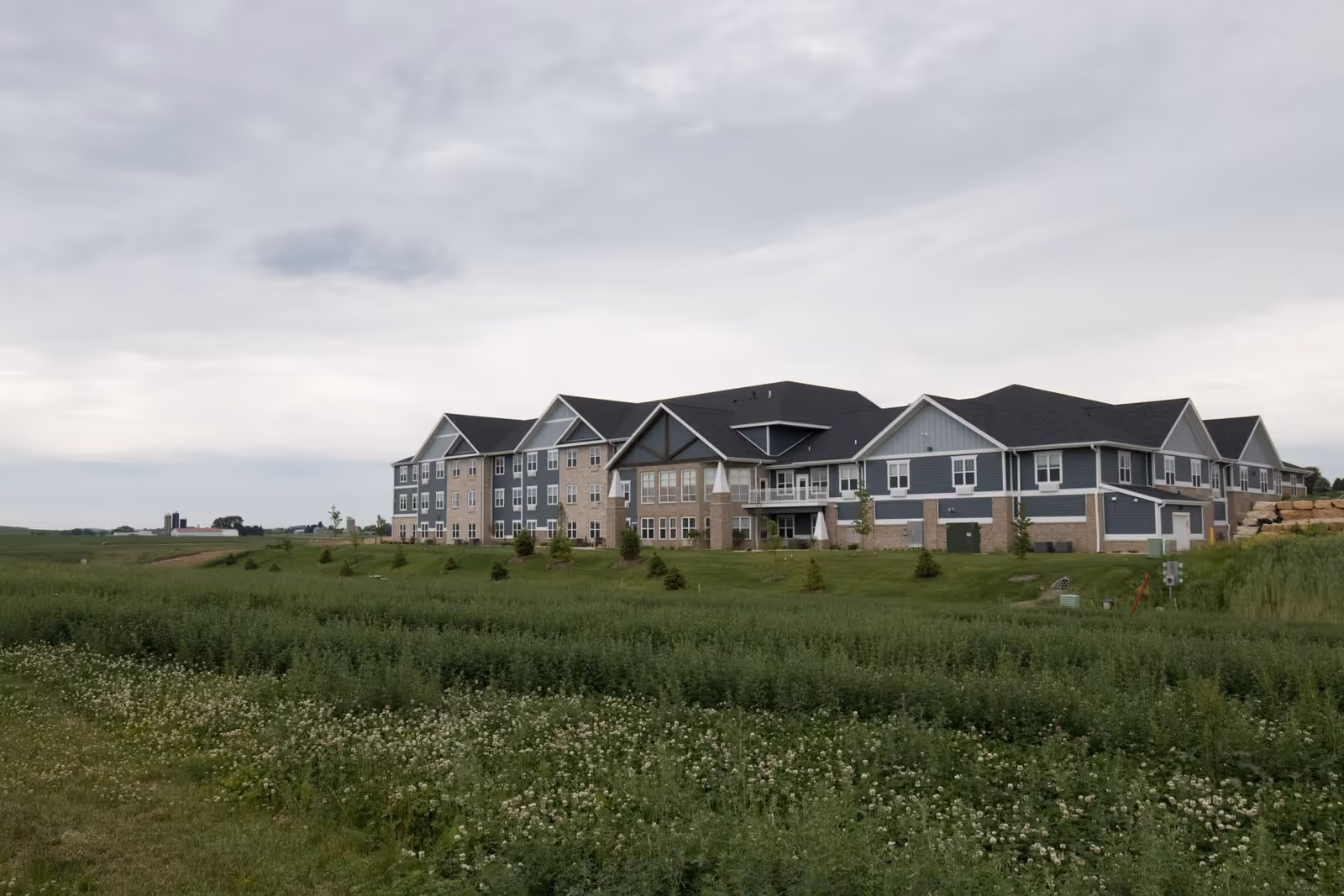 Exterior view of a large, multi-story assisted living and memory care facility building surrounded by green grass and small trees under a cloudy sky.