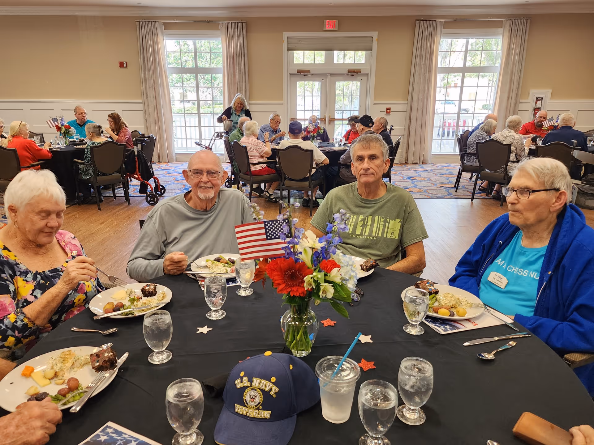 A group of elderly people sitting around a round table in a dining room, eating a meal. The table is decorated with a vase of red, white, and blue flowers and a small American flag. There are several glasses of water on the table, and a U.S. Navy veteran cap is placed in front. Other elderly people are visible dining at tables in the background near large windows and glass doors.