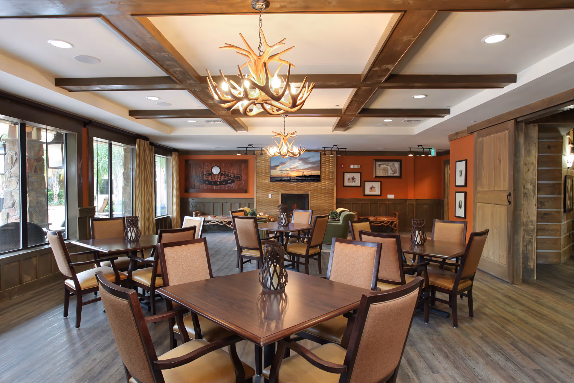 A cozy dining room with several wooden tables and cushioned chairs arranged neatly. The room features large windows on one side letting in natural light, wooden beams on the ceiling, and two antler-style chandeliers. The walls are decorated with framed pictures and a mounted TV above a fireplace. The flooring is wood, and there is a sliding wooden door on the right side.
