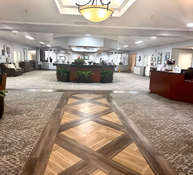 Spacious interior lobby area of a senior living facility with patterned carpet and wood inlay flooring leading to a central reception desk surrounded by plants. The ceiling features a large light fixture and the walls are decorated with framed pictures. Seating areas with chairs and sofas are visible on the left side.