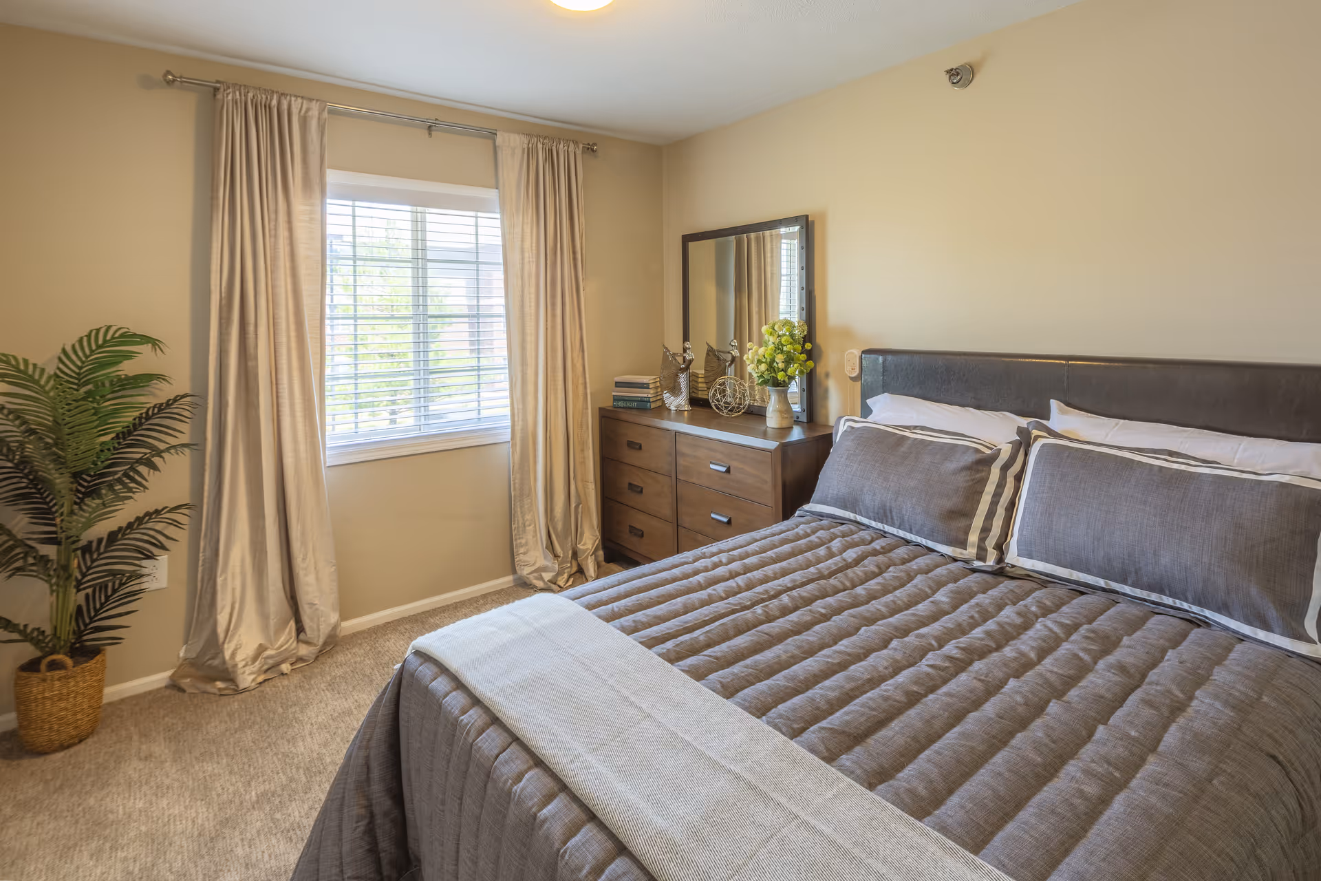 A bedroom with a neatly made bed featuring gray bedding and pillows. There is a wooden dresser with a mirror above it, decorated with books, a vase of flowers, and decorative items. A window with beige curtains allows natural light into the room, and a potted plant is placed in the corner.