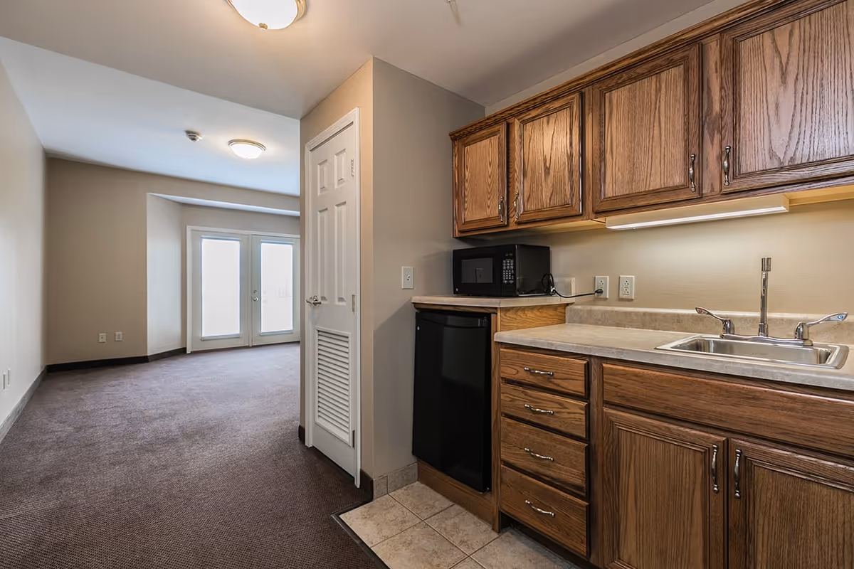 Interior of a senior apartment featuring a kitchenette with wooden cabinets, sink, microwave and mini-fridge opening onto a carpeted living area with double doors.