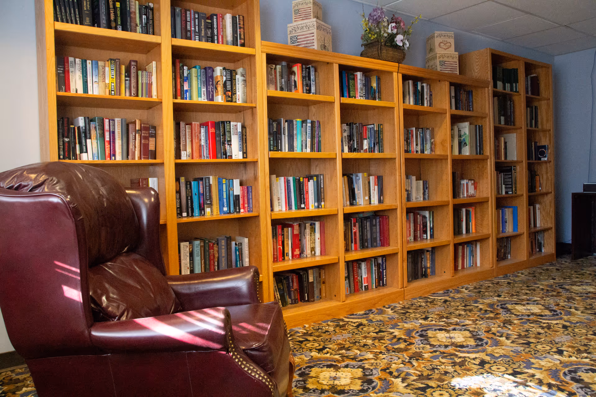 A cozy reading area featuring a large wooden bookshelf filled with books and a comfortable dark brown leather armchair on a patterned carpeted floor.