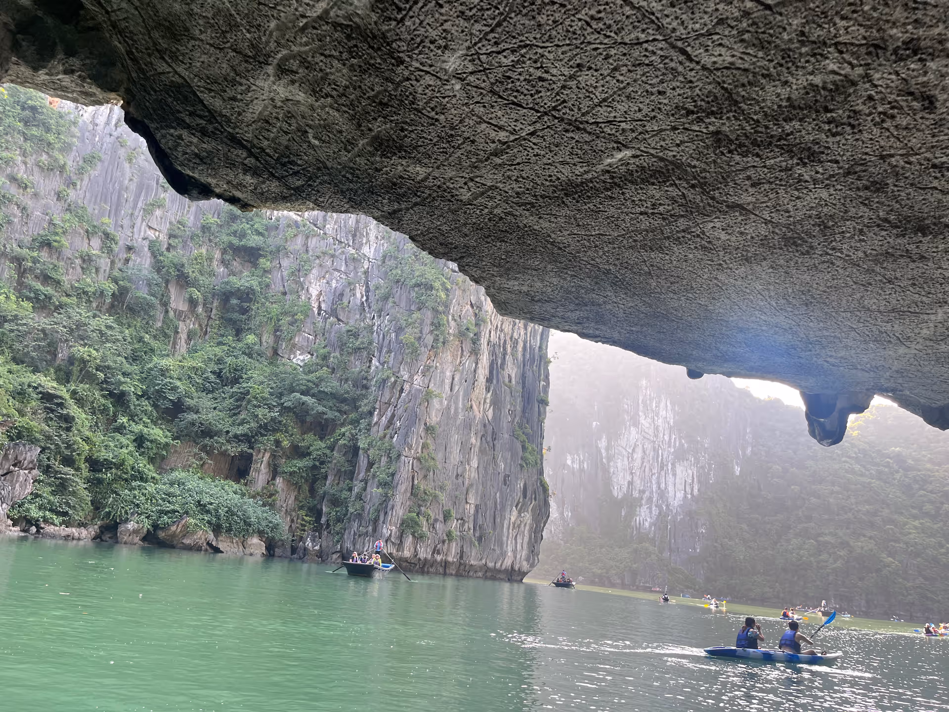 View from inside a cave looking out over a greenish body of water with several people kayaking and boating. Steep rocky cliffs covered with greenery surround the water.