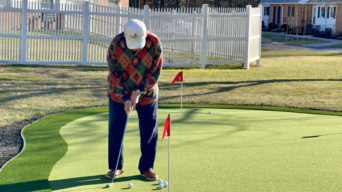 An elderly person wearing a white cap, colorful patterned sweater, and dark pants is putting on a small golf green with two red flags and several golf balls nearby. A white picket fence and houses are visible in the background under a clear sky.