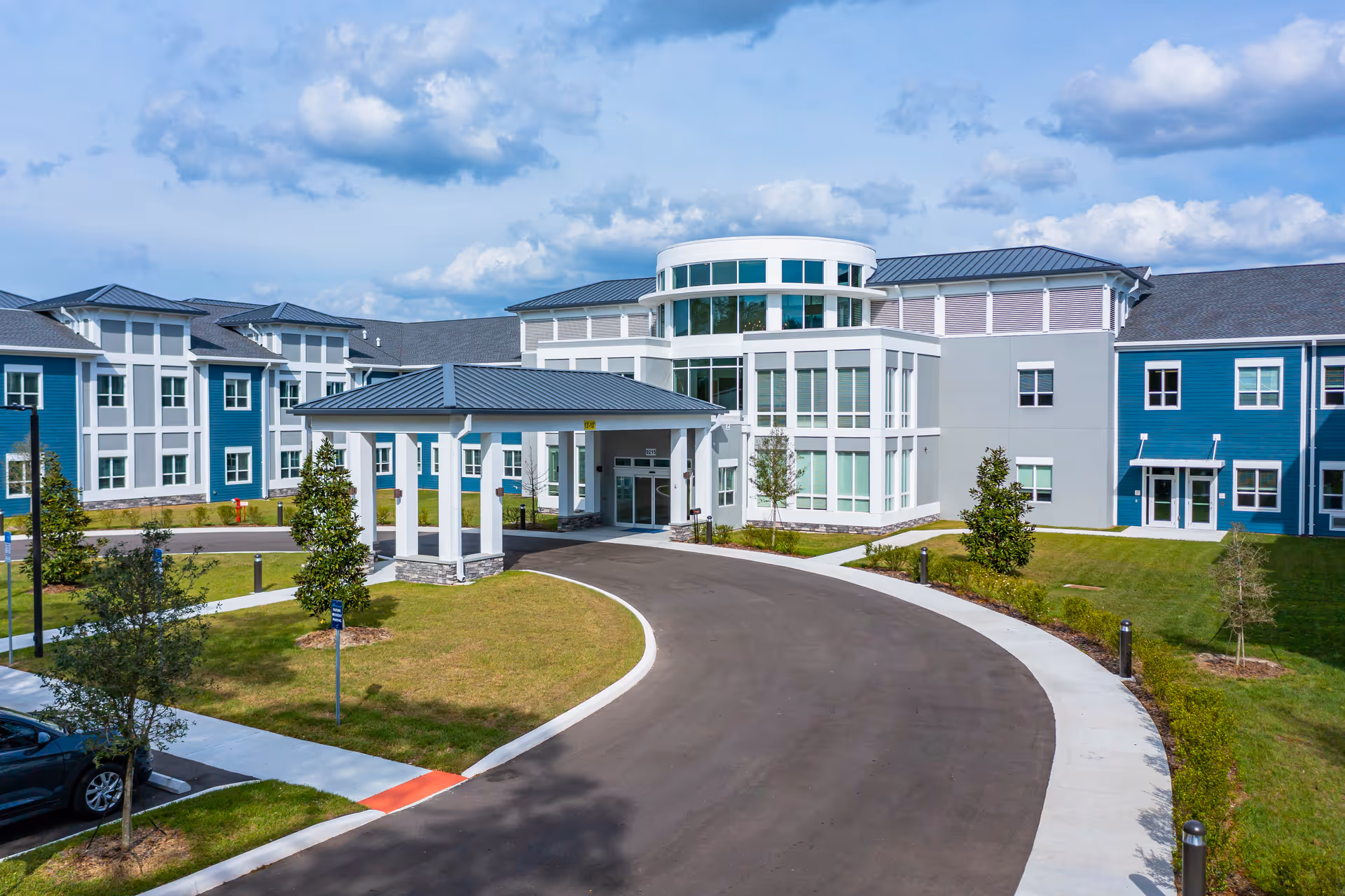 Front exterior of a modern three-story senior living building with a circular glass entry, covered porte-cochere, curved driveway, and landscaped grounds.