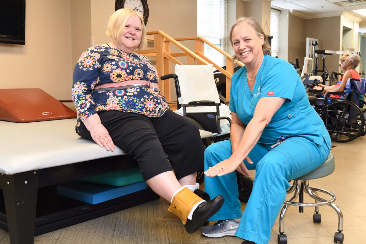 A senior woman sitting on a therapy table with an ankle weight on her leg, smiling at the camera. A healthcare professional in blue scrubs is seated on a stool next to her, also smiling and holding the woman's ankle. In the background, another elderly person in a wheelchair is visible in a rehabilitation room with exercise equipment.