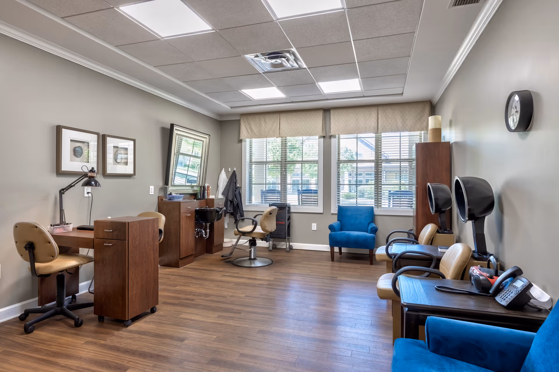 Bright salon-style common room with manicure stations, hair dryer chairs, and blue armchairs by large windows.