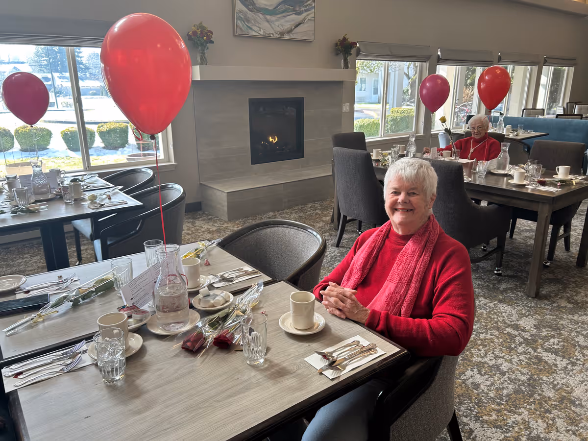 Two elderly women sitting at separate dining tables in a well-lit room with large windows and a fireplace. The tables are decorated with red balloons, roses, and place settings including cups, glasses, and utensils. Both women are smiling and wearing red clothing.