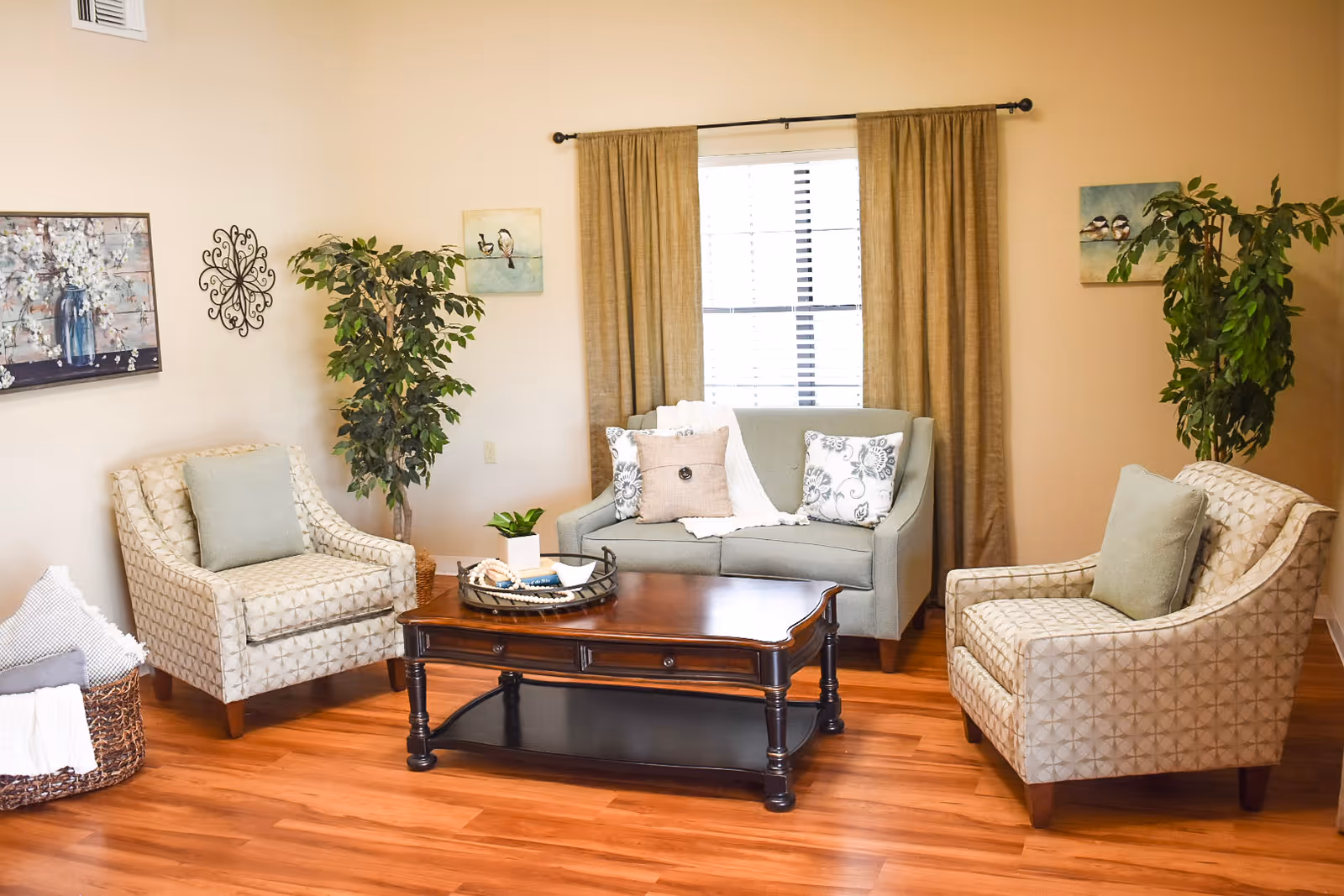 A cozy living room area with two patterned armchairs and a light gray loveseat arranged around a dark wooden coffee table. The room has wooden flooring, beige walls, two potted plants, and wall art including paintings and a decorative metal piece. A window with beige curtains is centered behind the loveseat.