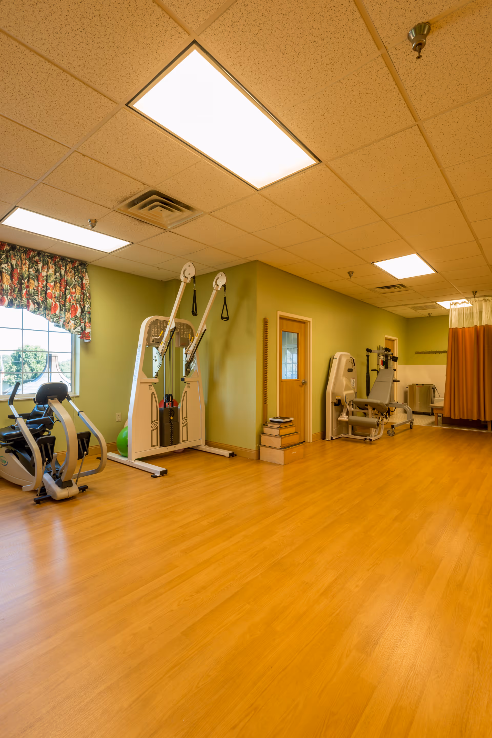 A spacious room with light wood flooring and green walls, equipped with exercise machines including a recumbent bike and resistance training equipment. The room has a window with floral curtains, fluorescent ceiling lights, and a wooden door. There is also a curtained area in the back corner.