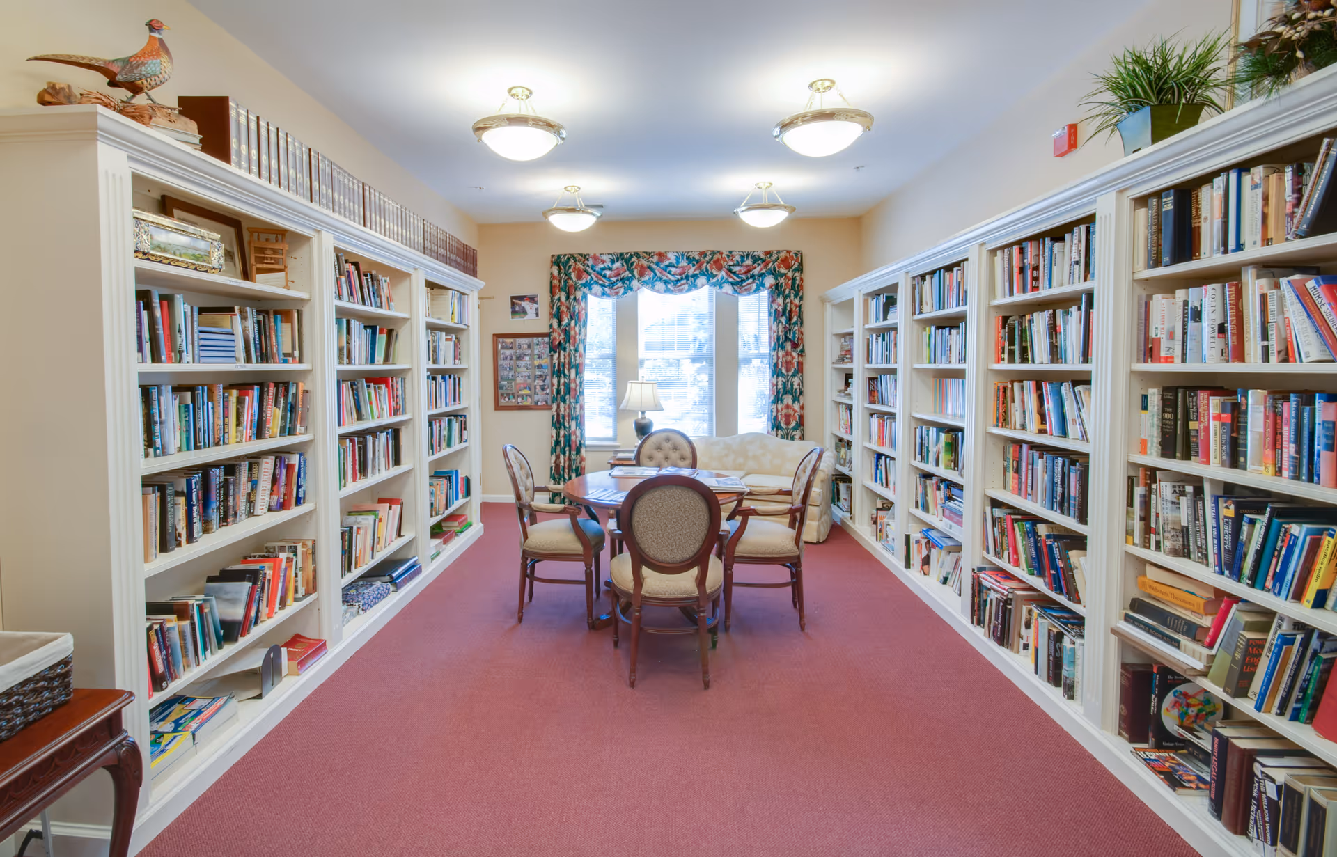 A cozy library room with white bookshelves filled with books lining both sides of the room. In the center, there is a round wooden table surrounded by four upholstered chairs. At the far end, a window with floral curtains lets in natural light, and a small lamp sits on a table in front of a cushioned bench. The room has a warm, inviting atmosphere with a red carpeted floor and ceiling lights.