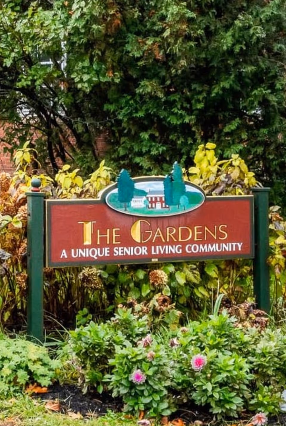 A wooden sign in a garden area surrounded by green plants and flowers. The sign reads 'The Gardens A Unique Senior Living Community' with an illustration of trees and a building above the text.