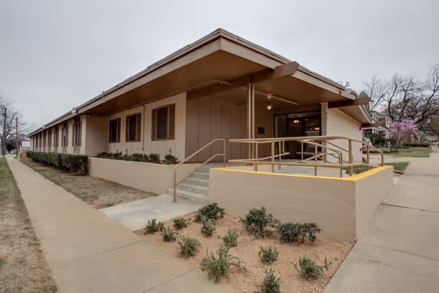 Front entrance of a single-story beige assisted living building with a covered entry, handrails, ramp, stairs and low landscaping.