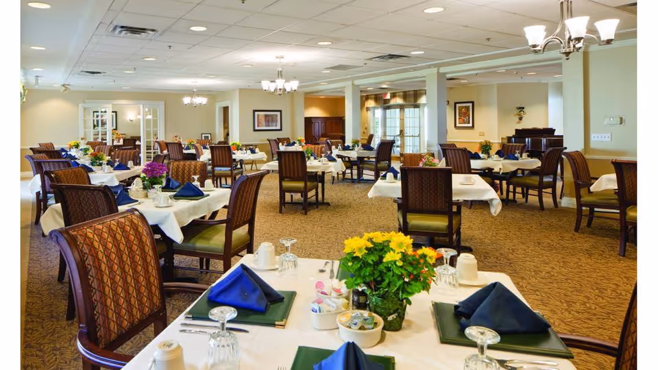 Bright senior living dining room with multiple tables set with napkins, glassware, and flower centerpieces.