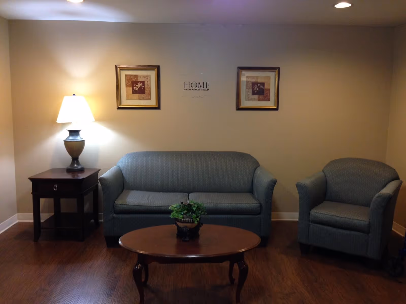 A cozy living room area with a blue upholstered sofa and matching armchair. A wooden coffee table with a small potted plant is in front of the sofa. To the left, there is a wooden side table with a lamp on it. Two framed floral pictures hang on the beige wall above the sofa, with a wall decal in the center that reads 'HOME WHERE MEMORIES BEGIN'. The floor is wooden and the room is softly lit.