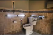 A bathroom with beige tiled walls and floor, featuring a white toilet with metal grab bars on both sides for accessibility. There is a toilet paper holder mounted on the wall to the left of the toilet and a call button below it. The upper part of the walls is painted a light brown color.