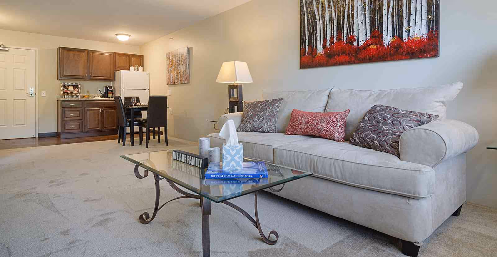 A cozy living area in a senior living facility featuring a light beige sofa with three decorative pillows, a glass coffee table with books and a tissue box, a floor lamp, and a colorful painting of birch trees with red foliage on the wall. In the background, there is a small kitchen area with wooden cabinets, a white refrigerator, a dining table with four black chairs, and a door.