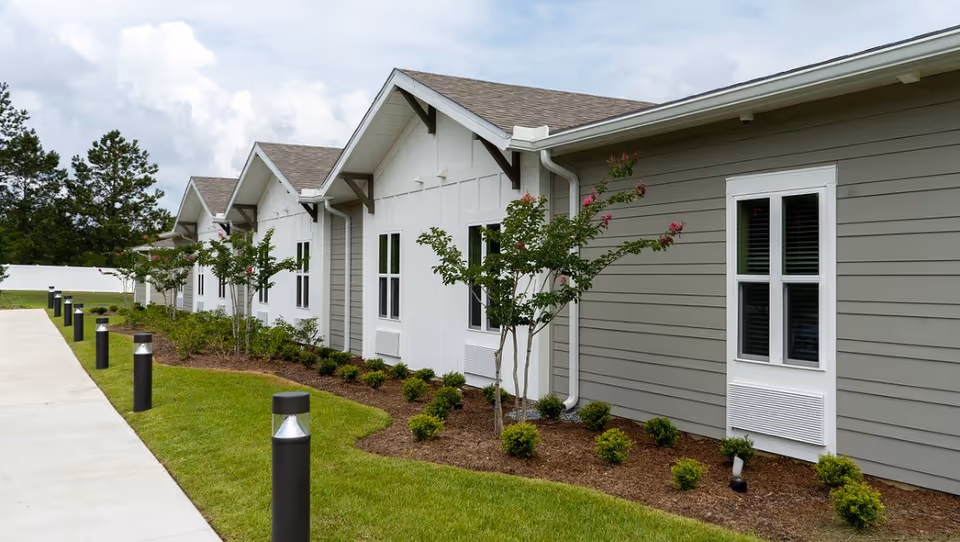 Exterior view of a single-story residential building with gray and white siding, multiple windows, small landscaped bushes and trees, and a concrete walkway with black bollard lights along the side.