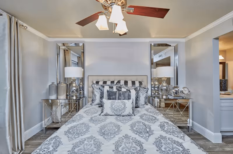 A neatly made bed with patterned bedding and multiple decorative pillows in a bedroom. The bed is flanked by two mirrored nightstands, each with a lamp and decorative items. The room has light gray walls, a ceiling fan with lights, and curtains on the left side.