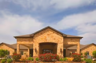 Front exterior view of a single-story building with a stone facade, a peaked roof, and a covered entrance. There are landscaped bushes and flowers in front of the building under a partly cloudy sky.