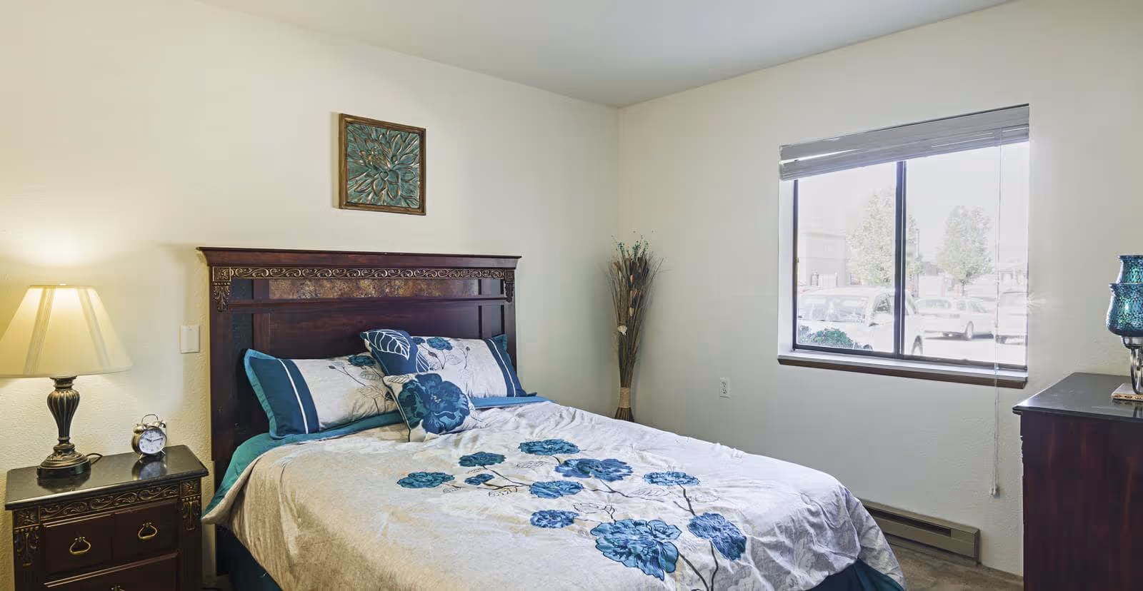 A bedroom with a bed featuring a dark wooden headboard and floral bedding in shades of blue and white. There is a nightstand with a lamp and an alarm clock on the left side of the bed. A window with blinds is on the right wall, letting in natural light. A decorative vase with dried plants is in the corner, and a framed floral artwork hangs above the bed.