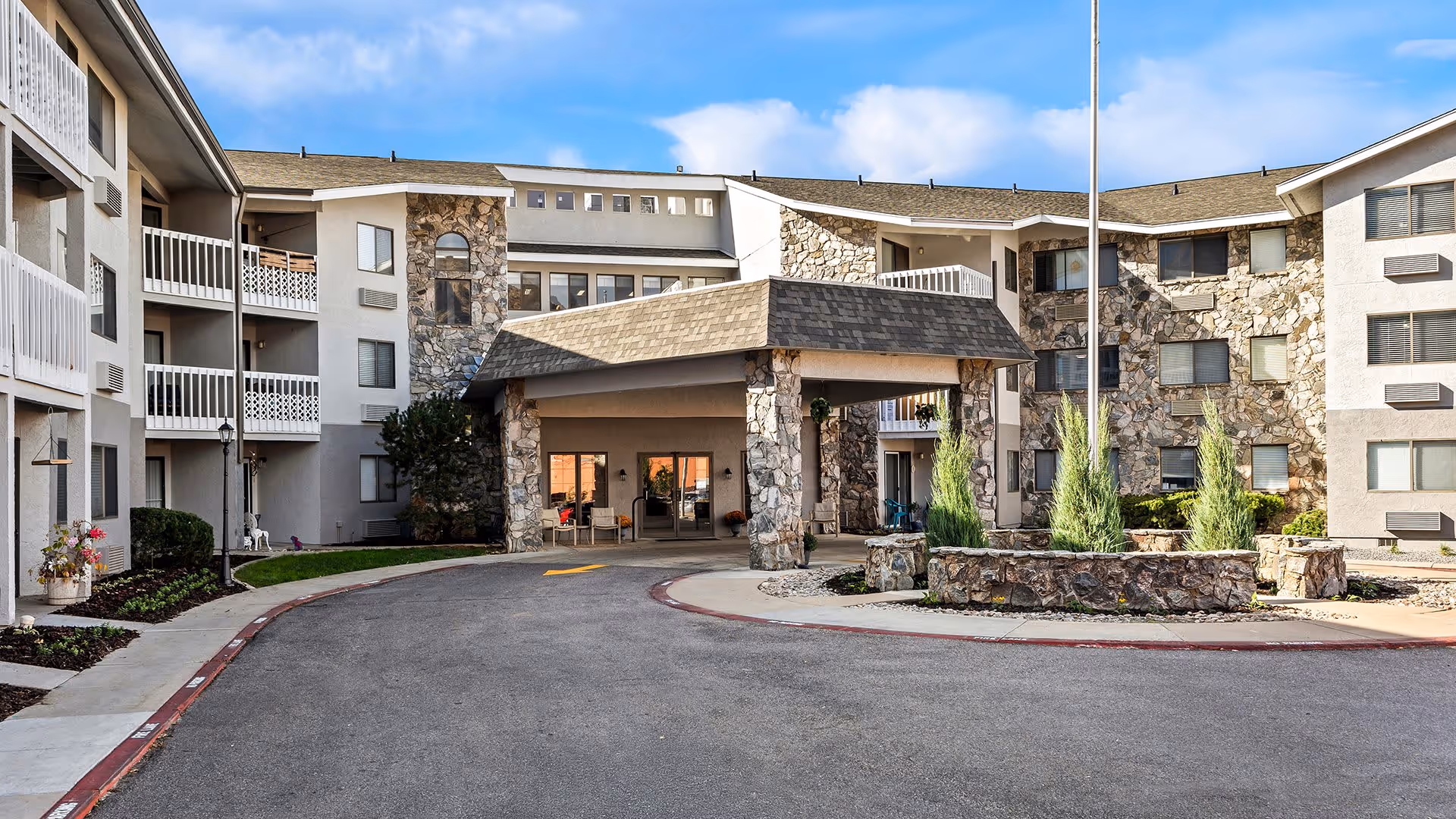 Exterior view of a multi-story retirement community building with stone and beige walls, balconies, and a covered entrance driveway. There are landscaped areas with plants and a flagpole in front of the entrance under a clear blue sky.