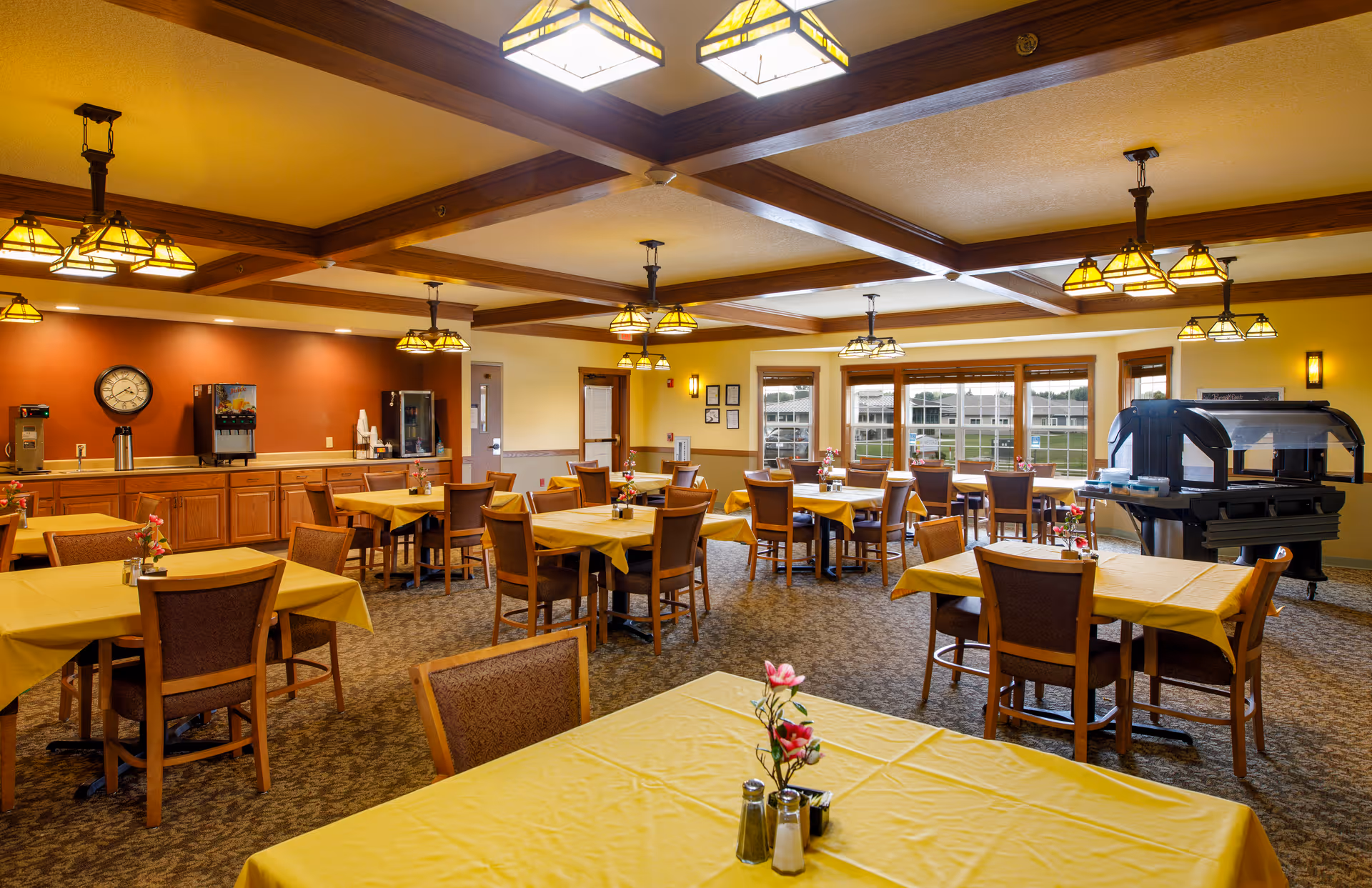 Well-lit dining room with multiple tables covered in yellow tablecloths, wooden chairs, pendant lights and a buffet/service area near large windows.