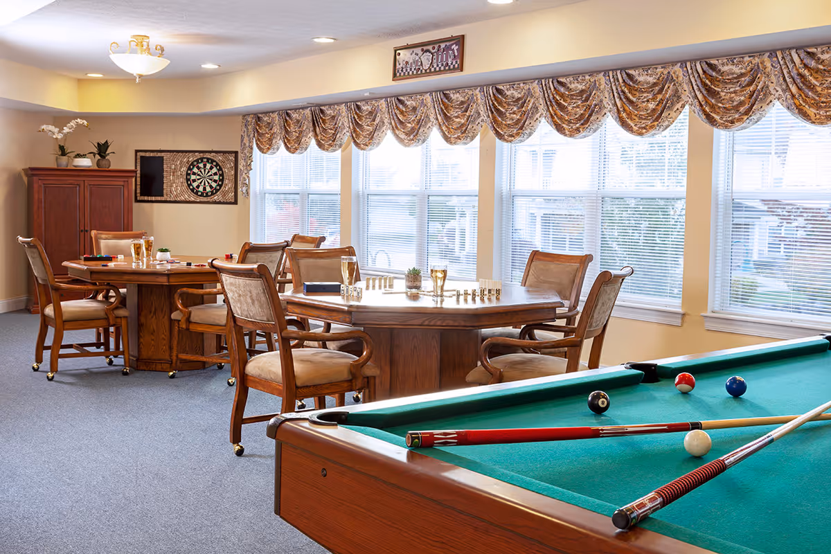 A recreational room with a pool table in the foreground, several wooden tables with chairs around them, and a dartboard on the wall. The room has large windows with patterned valances, allowing natural light to fill the space.