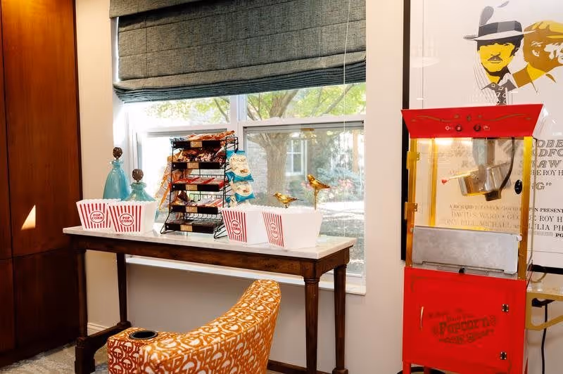 A cozy room corner with a wooden table holding popcorn containers, snack racks, and decorative bird figurines in front of a window with a gray roman shade. To the right, there is a vintage-style red popcorn machine next to a framed poster on the wall. An orange patterned chair is partially visible in the foreground.
