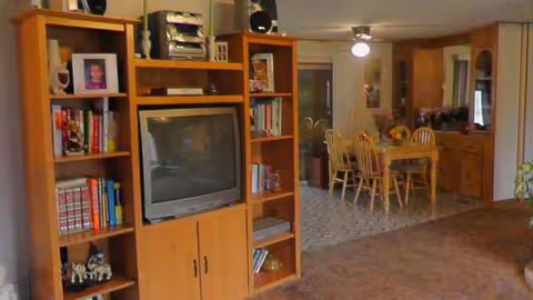 Wooden shelves with a TV and books, adjacent dining area with a wooden table and chairs.