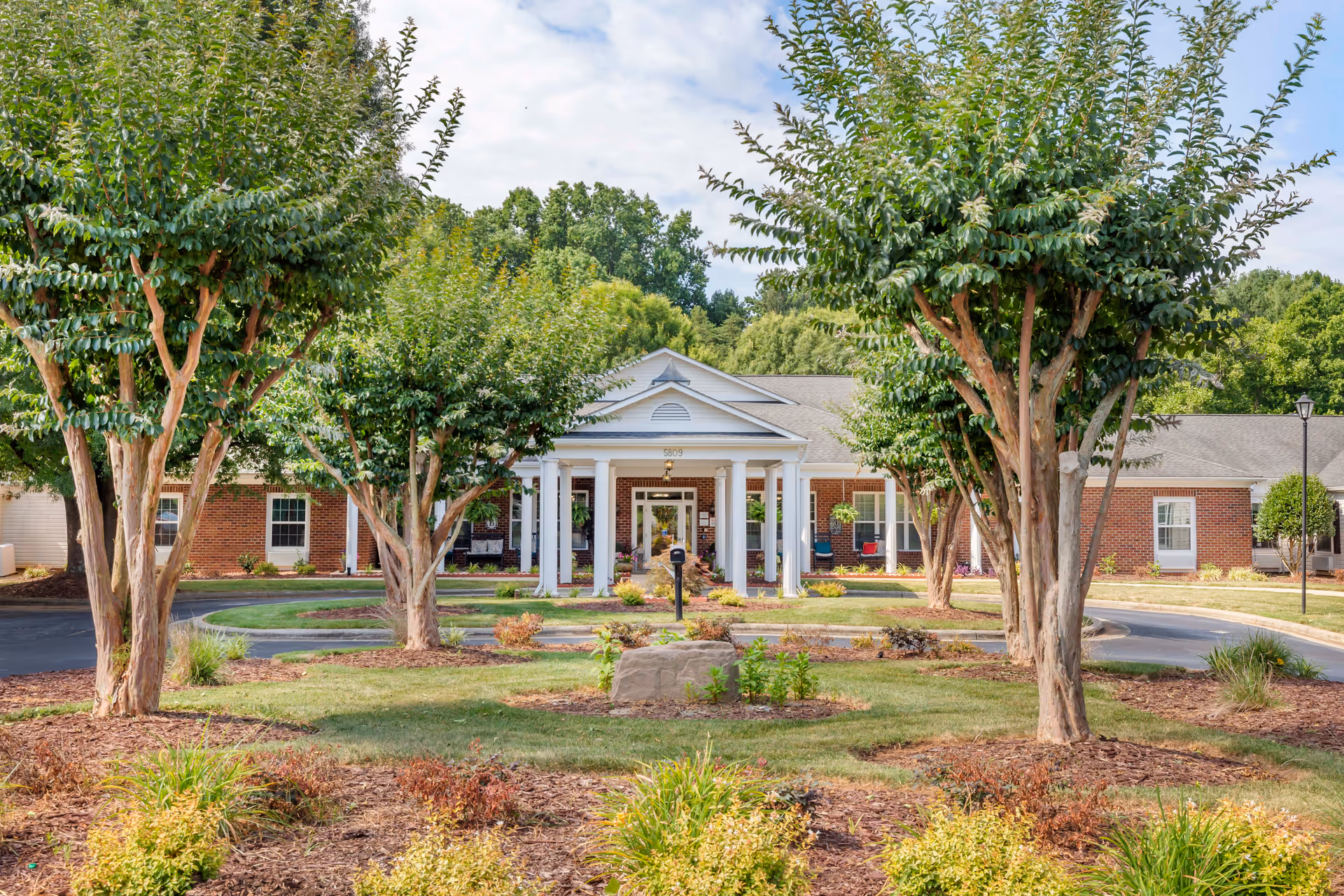 Front exterior view of a single-story brick building with white columns at the entrance, surrounded by landscaped greenery and trees under a partly cloudy sky.