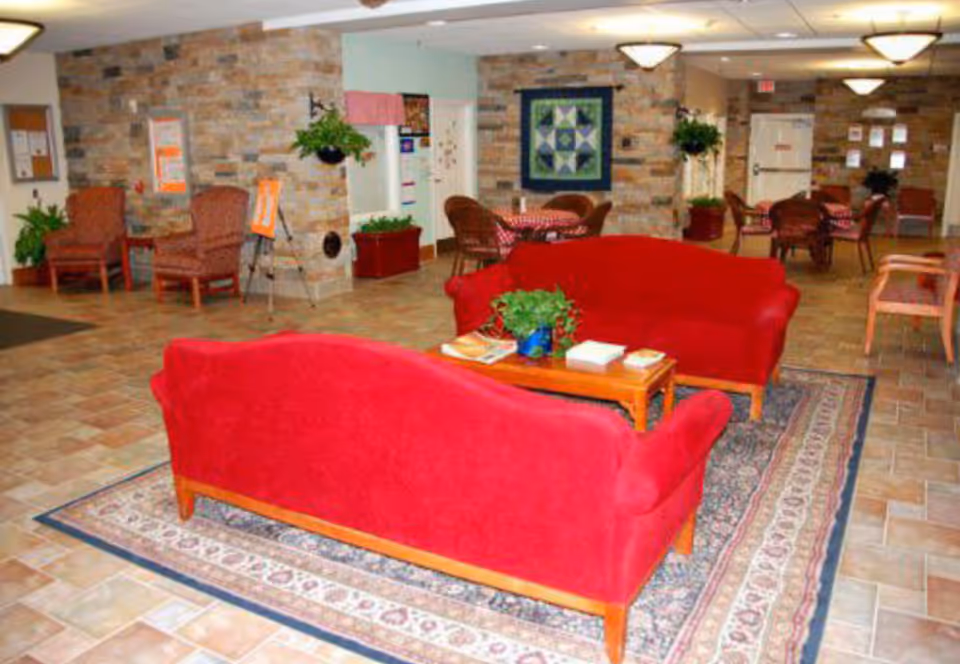 A senior living common area with two red sofas on a patterned rug, small tables, chairs and potted plants against stone accent walls.