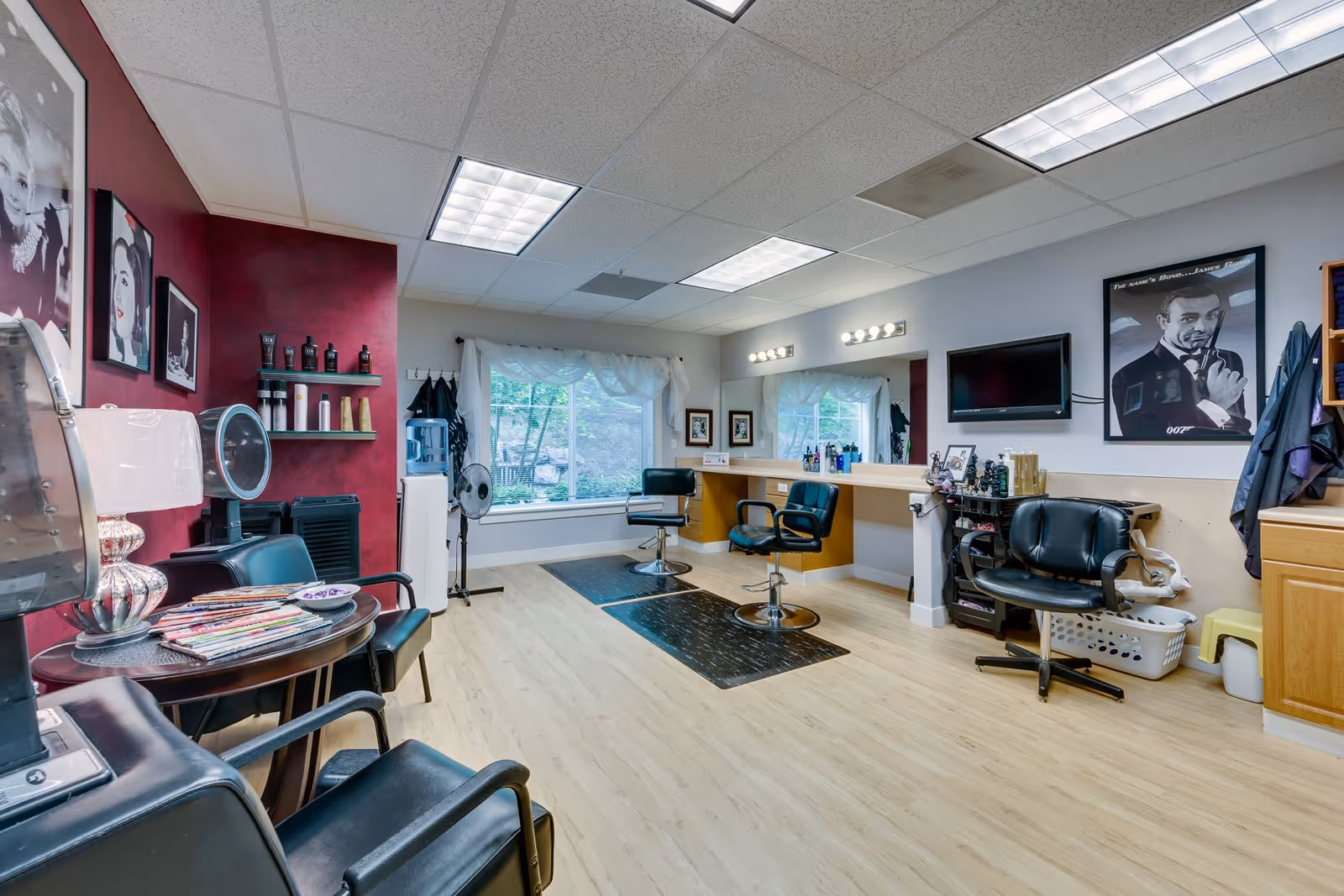 Interior of a salon area with styling chairs, mirrors, and hair care products. The room has light wood flooring, a large window with white curtains, and framed pictures on the walls including a James Bond poster. There are salon chairs, a small table with magazines, and various salon equipment visible.