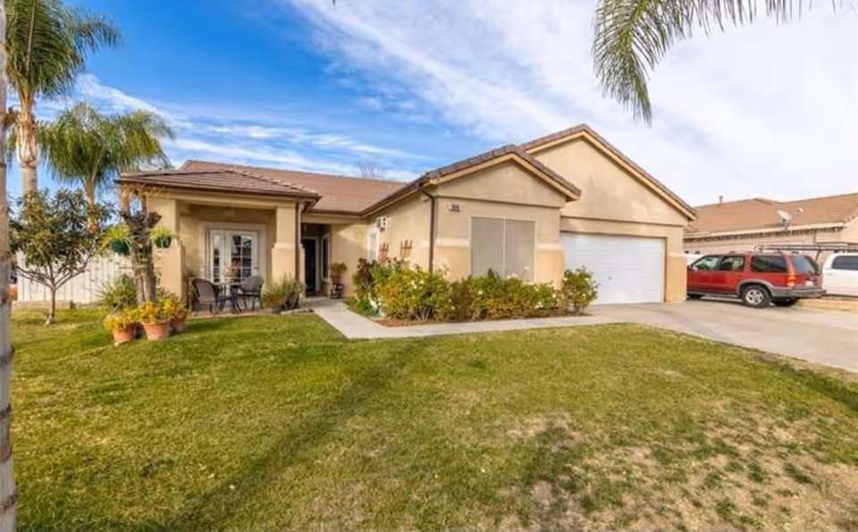 Front exterior of a single-story stucco house with a lawn, driveway, garage, and a small porch seating area.