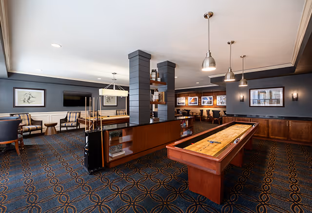 Spacious senior living common room with shuffleboard tables, seating areas, and framed artwork on the walls.