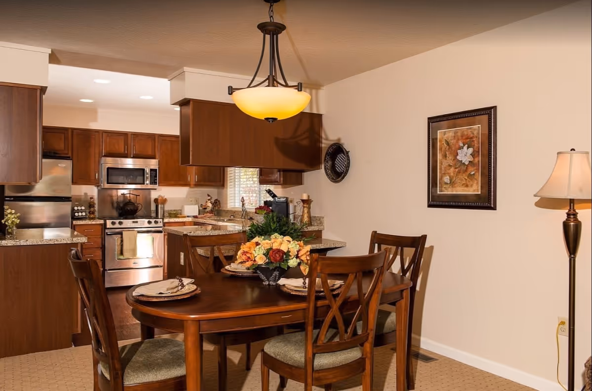A cozy dining area with a wooden table set for four, featuring a floral centerpiece. The dining area is adjacent to a kitchen with wooden cabinets, stainless steel appliances including a refrigerator, stove, and microwave. A hanging light fixture illuminates the dining table, and a framed floral painting and a floor lamp are visible on the beige walls.