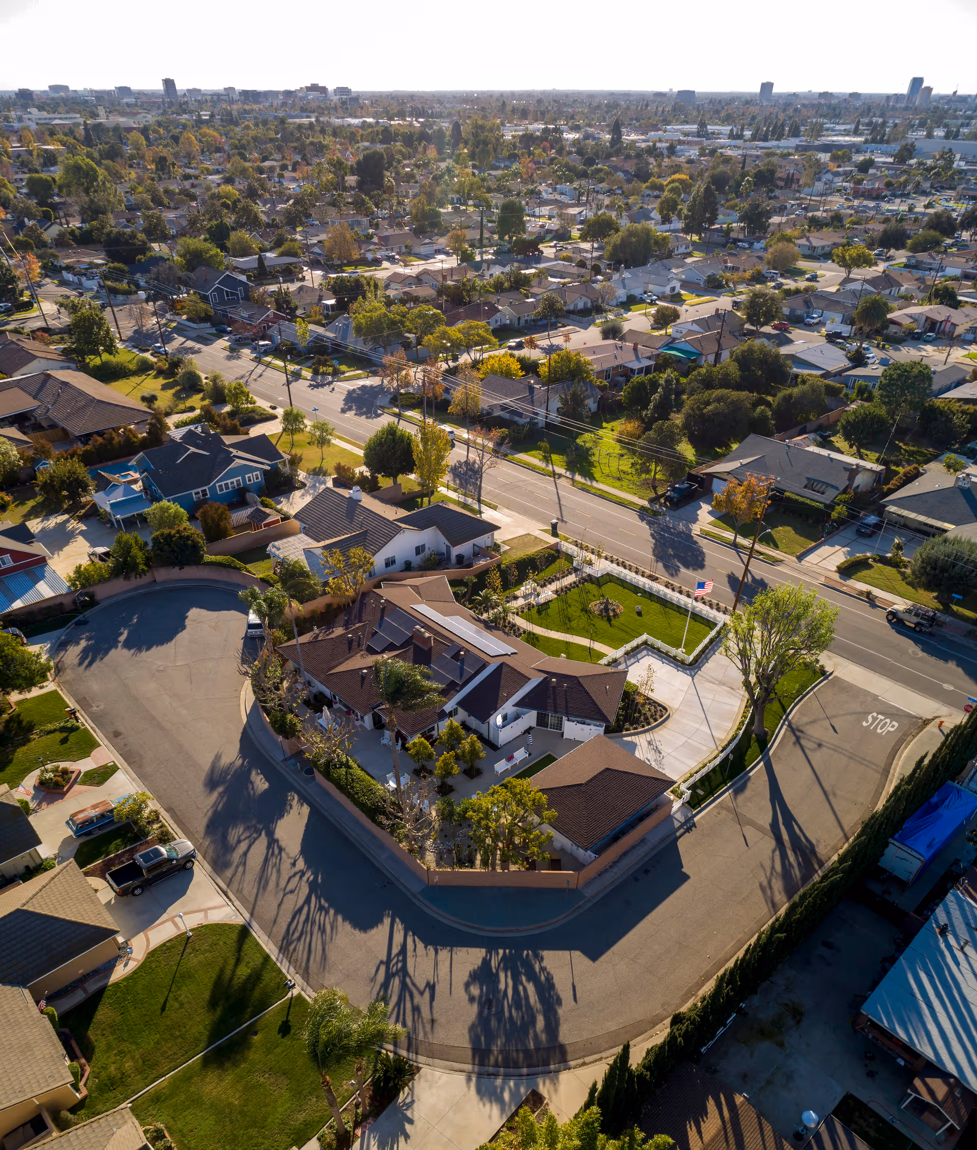 Aerial view of a residential neighborhood featuring a large house with solar panels on the roof, surrounded by a curved street and green lawns. The area includes multiple houses, trees, and a clear sky in the background.
