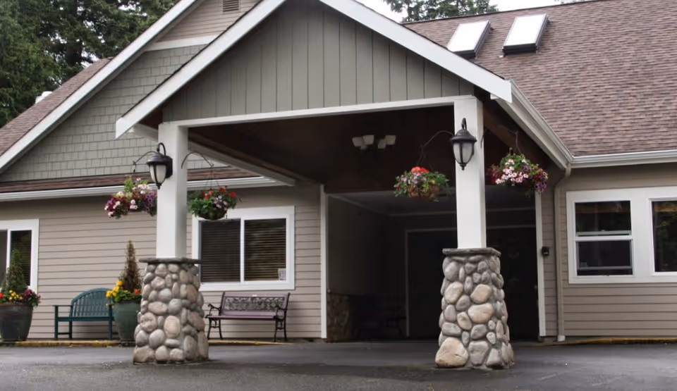 Covered entrance of a single-story building with stone pillars, hanging flower baskets, benches, and windows.